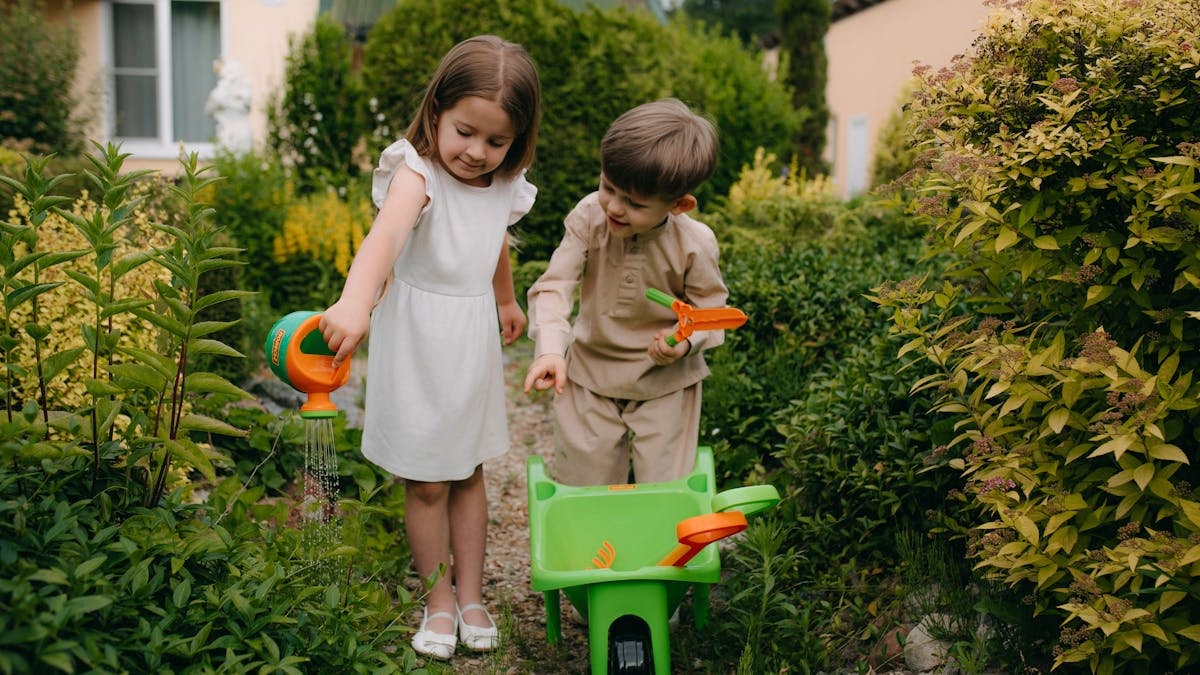 Two children playing in a lush garden, using a watering can and toy tools on a sunny day.
